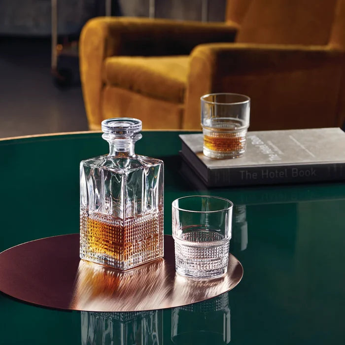 Novecento Whiskey decanter and glasses on a green table with a blurred chair in the background.